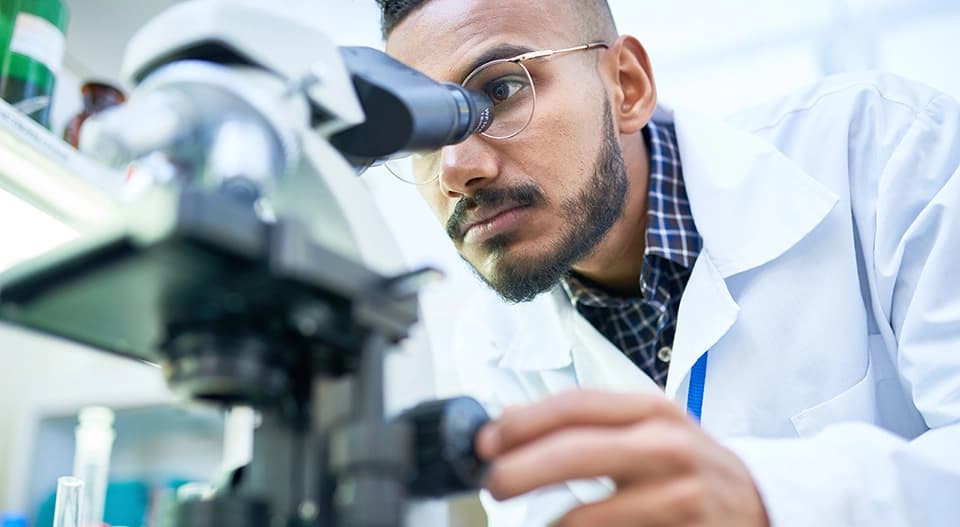 Lab technician in white coat examining sample through microscope in a medical or life science research setting
